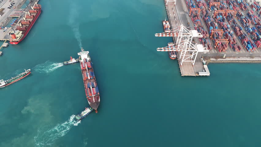 Large container ship being maneuvered by tugboats near a busy commercial port terminal. Concept of international logistics, maritime cargo transportation, and global trade infrastructure.