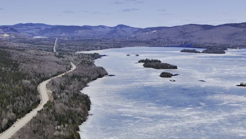 Aerial view of winding road along frozen lake in Quebec with forest and mountains under blue sky. g