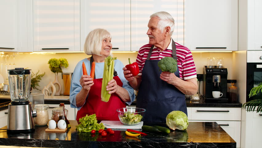 Senior old woman man smiling in home kitchen selecting fresh vegetables for healthy fit diet meal. Elderly grandmother grandfather share love while cooking together for wellness and family nutrition.