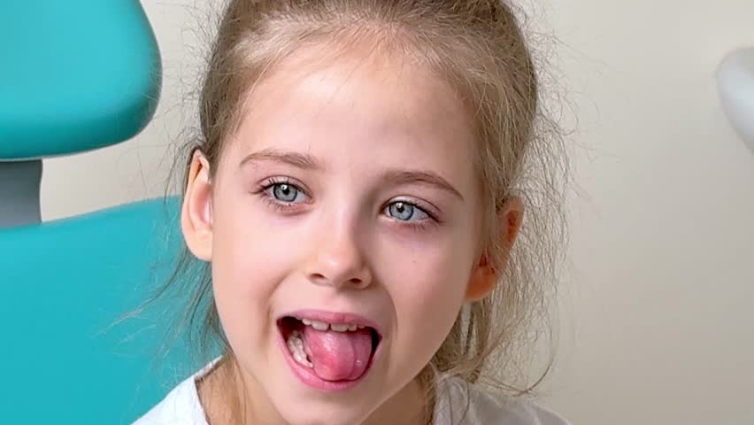 Little girl with light brown hair and blue eyes sits in a dental chair, sticking out her tongue during a dental examination, with dental tools and equipment visible in the background