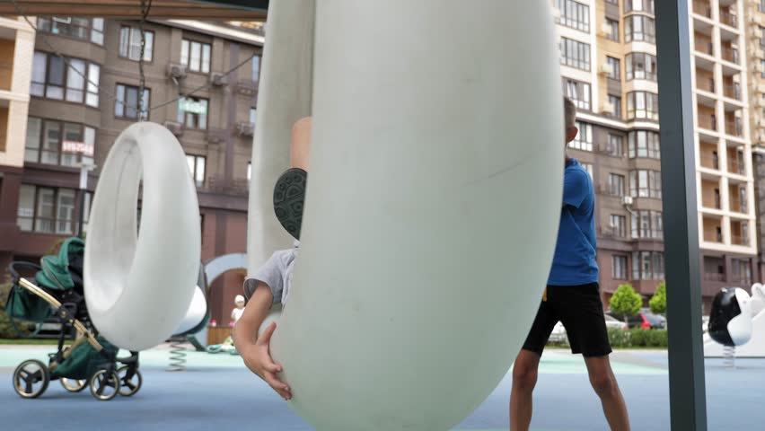 A happy boy is lying on a modern round plastic swing in a modern courtyard, against the background of a new high-rise residential building. In summer, children walk in the courtyard.