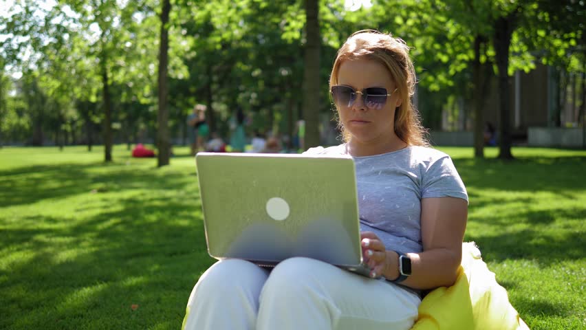 A young woman with a laptop is sitting in an easy chair in a modern park. The camera is stabilized, and the wind is blowing her hair. A freelance opens her laptop to take pictures of the park.