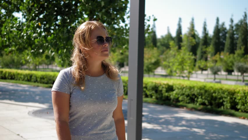 A young woman with blonde hair, wearing sunglasses, walks through a modern city park on a summer day in sunny, hot weather. The girl walks slowly and looks into the camera.