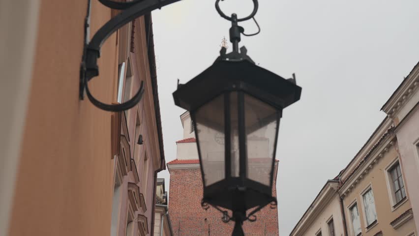 Krakow. Narrow street lined with historic buildings, viewed from a low angle. In the foreground, a black metal street lantern hangs from a curved wall-mounted bracket. The lantern is slightly out of focus, creating a soft silhouette against the lighter background. Behind it, the street is framed by warm-toned facades beige and light brown buildings with traditional architectural details such as rectangular windows and decorative trim. In the center background, a taller structure made of red brick rises above the surrounding buildings. It features a clock face and a small tower or steeple at the top, suggesting it may be part of a church or historic landmark. The sky is overcast and pale gray, giving the scene a muted, calm atmosphere. The overall composition emphasizes depth, with the blurred lantern in the foreground and the sharper architectural elements receding into the background, evoking a quiet, old-town European street setting.