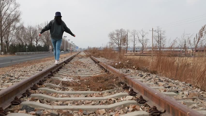 Cinematic stock video of a woman walking freely along rural railway tracks. The footage captures the female figure moving forward on railroad rails, with arms outstretched, set against an overcast sky, open countryside, and roadside landscape. High-quality footage exploring themes of freedom, journey, solitude, and wanderlust, ideal for lifestyle, travel, and adventure content.