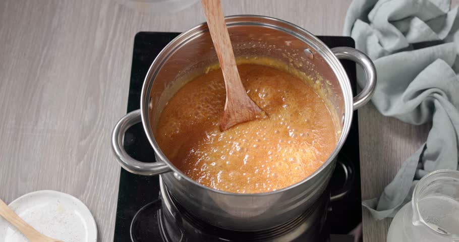 Close up of hot homemade caramel sauce boiling in a pot on a stove while salt is added, creating salted caramel on a gray kitchen table. Rich and glossy sauce made from sugar, butter, cream and salt. Cooking process concept