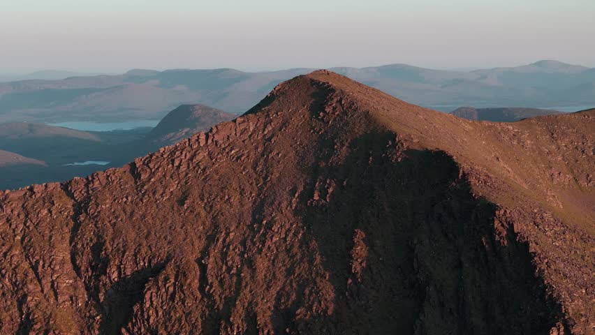 Aerial view captures the majestic mountain range. Rugged mountain peaks against a clear sky. Sunrise illuminates the textured mountain slopes.