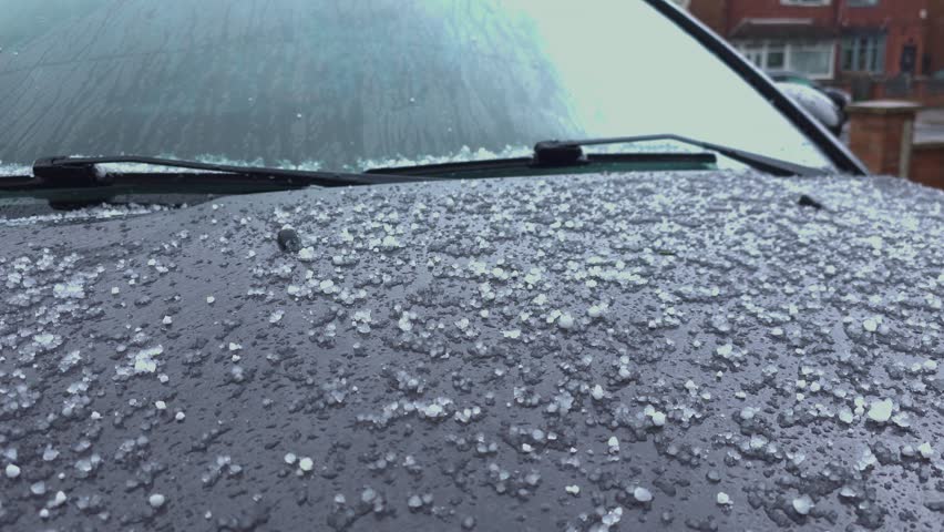 Hailstorm hitting parked car with ice pellets covering windshield and hood in residential area. Extreme weather conditions increasing storm damage risk with frozen precipitation on urban street