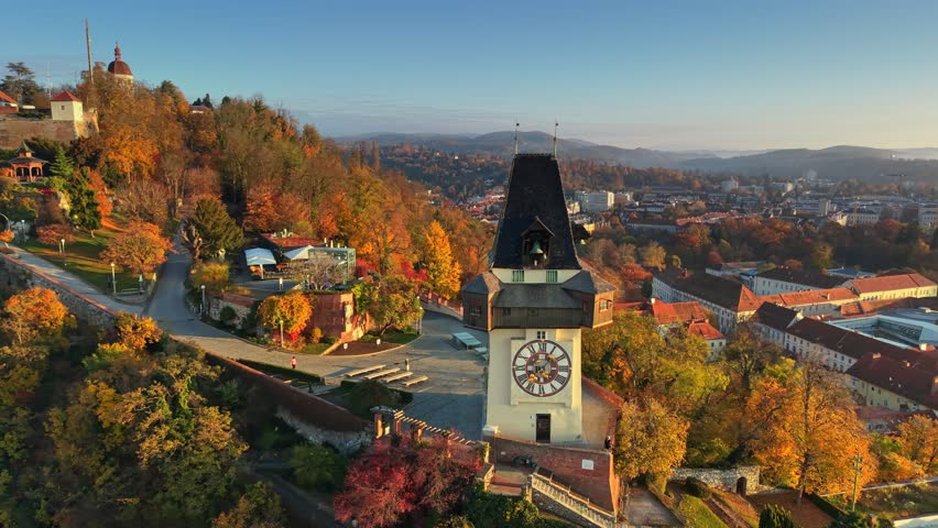 Aerial view of iconic Clock Tower in Graz, Austria. Famous Grazer Uhrturm on Schlossberg hill is surrounded by vibrant orange trees and panorama of Styria