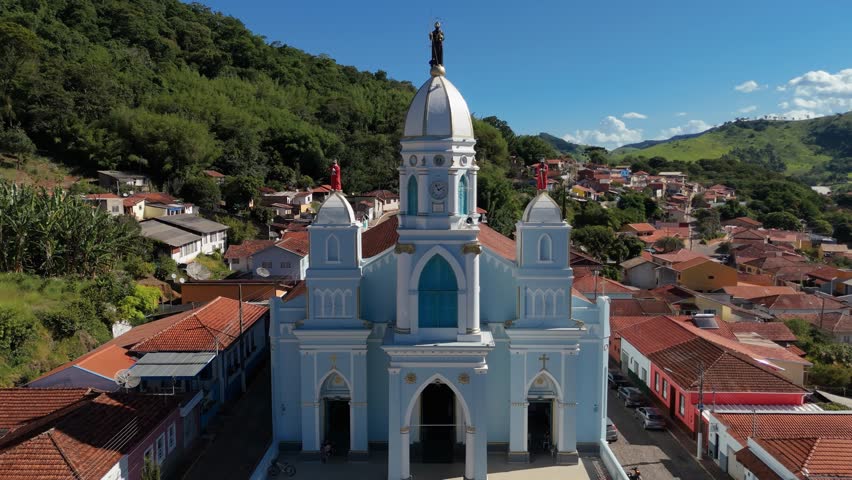 Aerial view of blue and white Matriz Church in town square surrounded by red roof houses and green mountains in Sao Bento do Sapucai, Brazil. Pull back and pan revealing town
