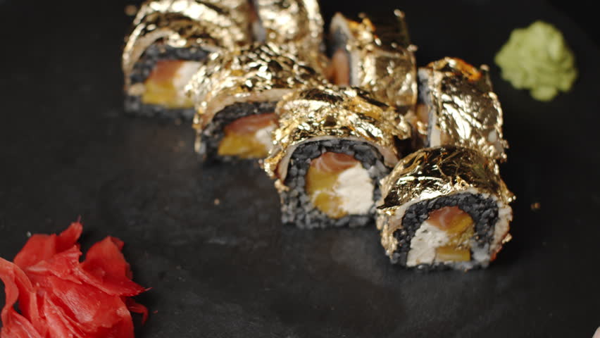 Luxurious sushi roll covered with edible gold leaf rotating on black slate plate with wasabi and pickled ginger presenting a delicacy from asian cuisine and fine dining