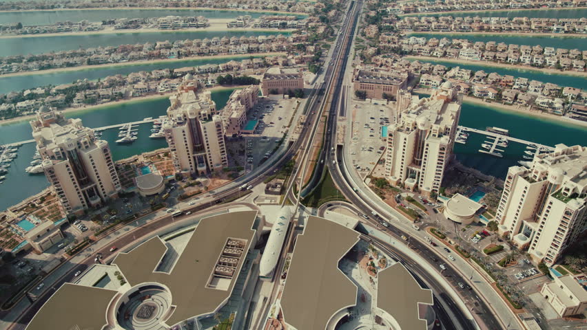 Aerial pan view of Palm Jumeirah island showing a busy road with buildings on both sides and water with boats nearby.
