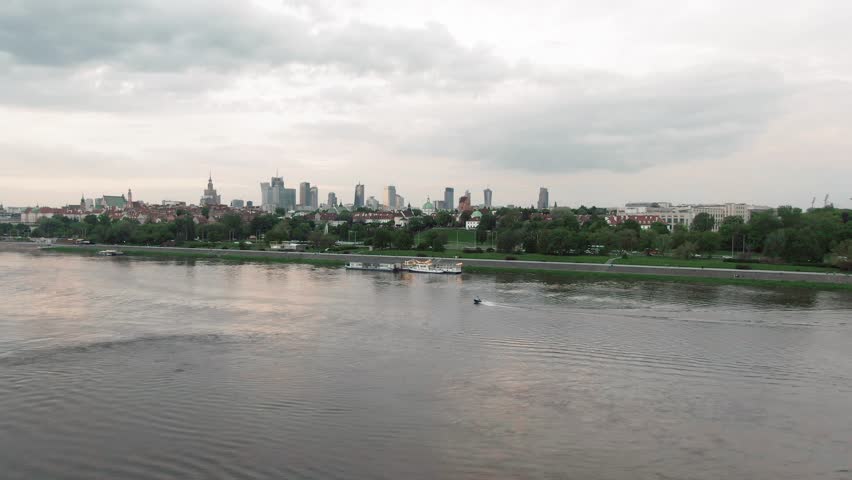 High angle wide shot of the Vistula River with a small boat traveling past the Warsaw city skyline under an overcast sky