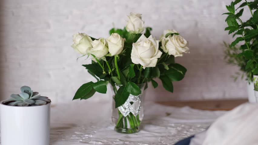 Beautiful bouquet of fresh white roses in a glass vase decorated with silver hearts on a table