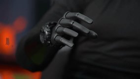 Close-up of a robotic or prosthetic hand resting against a person’s torso. The hand appears to be made of dark, matte material with articulated fingers, designed to mimic human movement. Each finger segment is clearly defined, giving it a mechanical yet lifelike structure. The wrist area includes a metallic joint or casing, suggesting advanced engineering or wearable technology. The hand is gently touching or positioned near a dark fabric surface, likely clothing. The background is softly blurred with warm orange lighting on one side, creating a contrast with the cool, dark tones of the robotic hand. The overall mood feels futuristic and slightly dramatic, emphasizing themes of technology, robotics, and human-machine interaction. - Powered by Shutterstock - Get 15% off with code: PIKWIZARD15