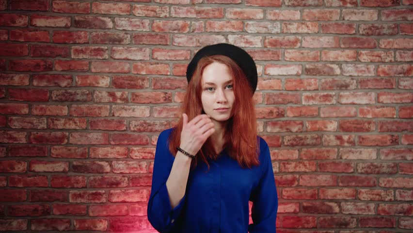 A portrait of a dreaming joyful young caucasian redhead woman in a blue blouse and black beret, fantasizes and thinks about pleasant good things against a brick wall. She twirls a hair and looks up