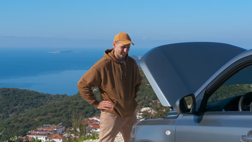 Stressed man standing by broken down car. Frustrated young man looking at his broken car with an open hood on a scenic mountain road overlooking the sea, waiting for assistance during a road trip