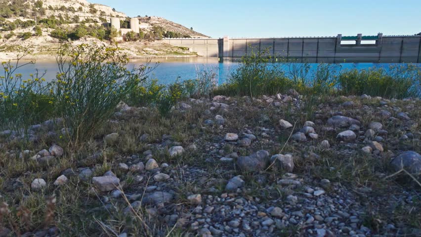 4k drone footage captures a dramatic take off from a rocky hill adorned with yellow flowers. The camera rises to reveal the complex engineering of Amadorio dam wall, showing the crest, spillway, and gate control house in detail. The upstream water level and free board are clearly visible against the calm reservoir. Set against a vast blue sky, this technical yet scenic shot highlights infrastructure and nature in harmony. Ideal for engineering and travel projects