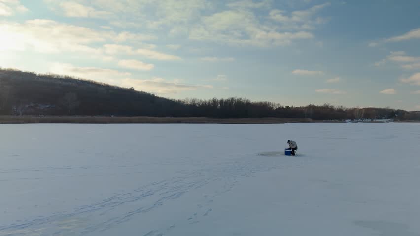 Individual dressed in winter clothing fishes on frozen lake surface, with distant hills and a bright sunset illuminating the serene winter landscape