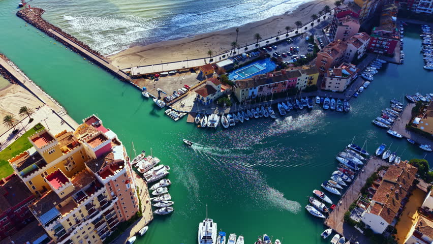 Bright boats are docked in a marina near a small coastal town in Spain. People can be seen enjoying the beach while waves crash nearby. The scene shows a clear day with some trees.