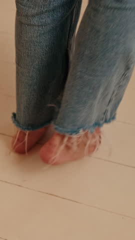A woman in jeans walks barefoot on a light wooden floor in a kitchen. She wiggles her feet, showing dark nail polish.