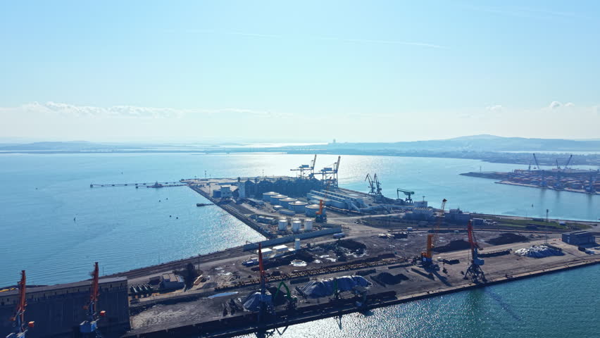 A large industrial port shows cranes, ships, and storage buildings along the coastline. Water reflects the sky, creating a lively scene during daytime hours.