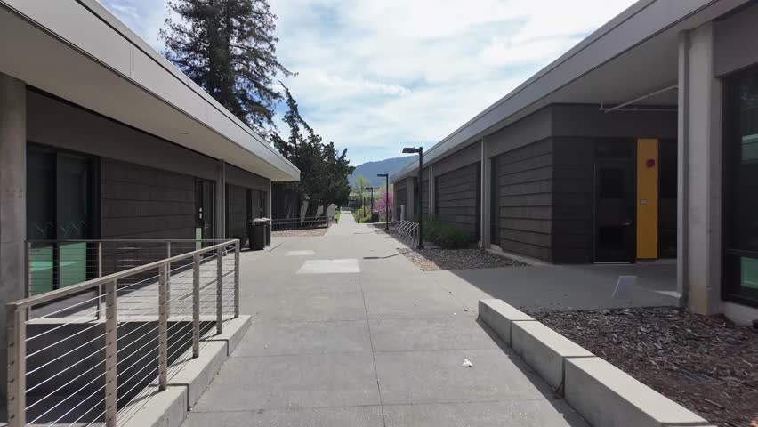 First-person perspective walking down a sidewalk between modern buildings and manicured landscaping at West Valley College campus in Saratoga, California on a sunny day with blue sky