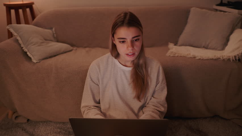 Young woman sitting comfortably at home, deeply focused on her laptop screen. She is engaged in online work or study in the cozy ambiance of her living room during the evening hours.