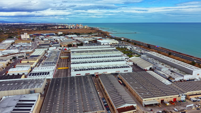 A wide view of a coastal industrial area with several factories visible. The city skyline is in the distance. Water and land contrast in this location on a clear day.
