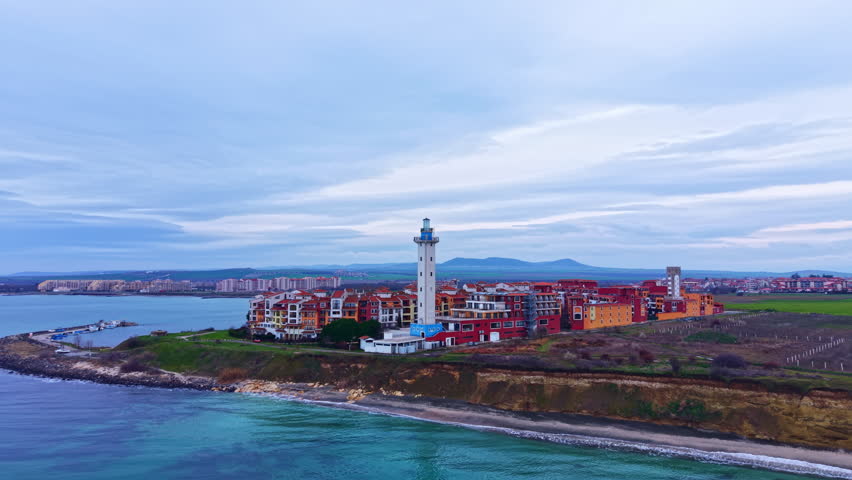 A coastal scene shows a lighthouse standing tall near a town with buildings along the shore. The ocean is visible with waves and the sky has clouds, creating a typical day at the coast.