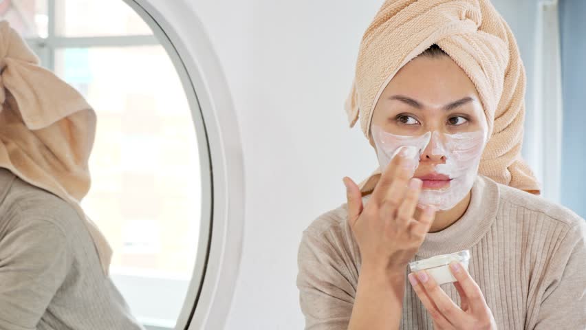 Asian woman with towel wrapped around her head looking at mirror and smoothing cream over face.