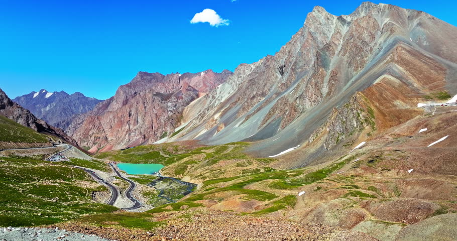 Dramatic aerial view of high mountain peaks and turquoise lake along a winding mountain pass road in Xinjiang, China.