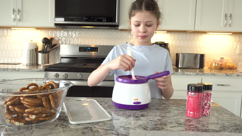 With focused attention, this budding culinary artist dips pretzels into a pot of melted chocolate, creating sweet delights in the warmth of a well-lit home kitchen.