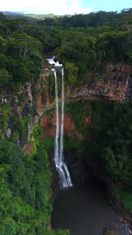 High-angle view shows waterfall plunging into deep pool. Water cascades down rocky cliffs into calm pool below. Dense forest surrounds dramatic canyon landscape