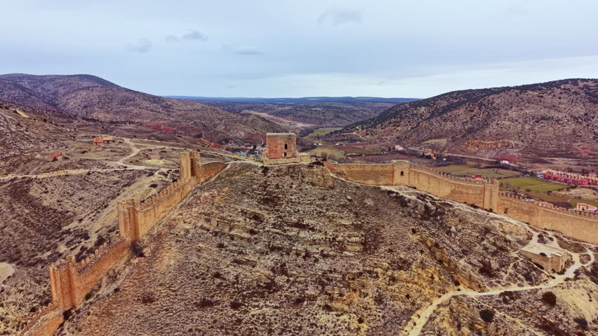 A large castle sits on top of a hill. The landscape shows mountains and valleys. The sky is gray and cloudy above. Fields can be seen in the background.