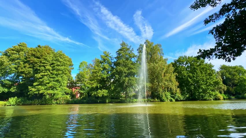 A tranquil park scene unfolds with a vibrant blue sky and fluffy clouds overhead. A majestic fountain sprays water over a calm pond surrounded by rich greenery.