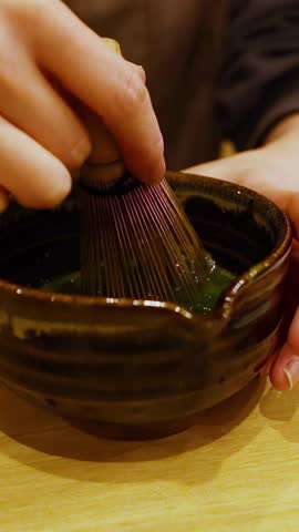 Close-up shot of hands using a bamboo whisk to mix matcha powder in ceramic bowl