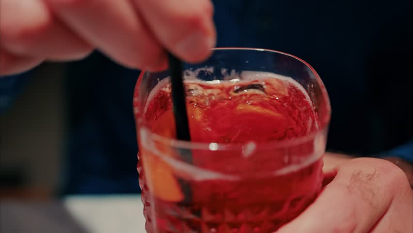 A person stirs a vibrant red cocktail with ice and orange slices using a straw in slow motion.