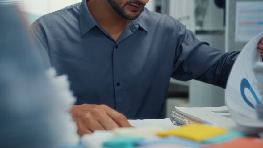 Closeup overworked young Latin businessman reading document in large stack of paperwork file at office desk. Tax audit deadline, accounting stress, corporate burnout, bookkeeping, bureaucracy concept.