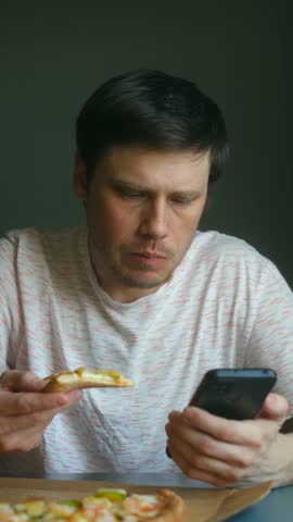 Adult man enjoying slice of pizza for lunch while simultaneously browsing social media or scrolling through his phone, distracted from his meal in modern concept of eating habits. Vertical.