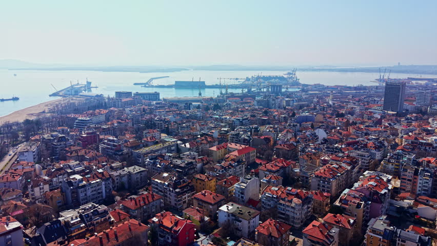Aerial view captures many buildings in city next to harbor. The area shows a mix of houses and commercial sites under clear sky. Water and docks are visible in distance.