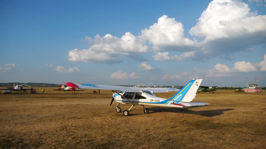 Small aircraft taxiing across an open airfield, showcasing the gradual movement and preparation for takeoff, surrounded by various vintage planes and a vibrant sky with fluffy clouds