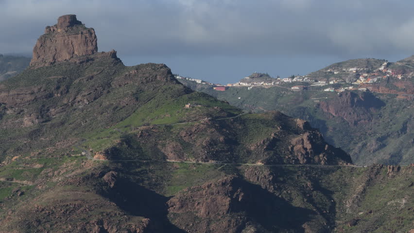 Panoramic view of a rugged volcanic mountain landscape in Gran Canaria, featuring a winding road, distant village and dramatic, moody cloudy sky.