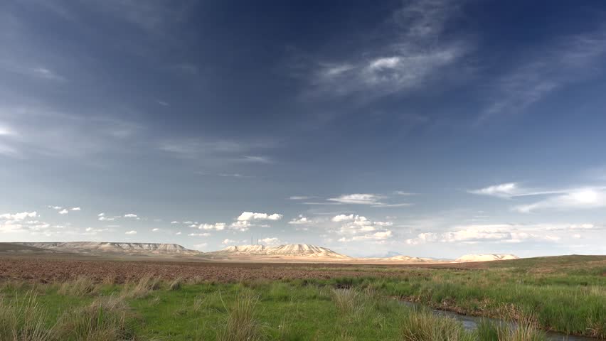Time-lapse view of Turkmenistan’s vast barren plains and dry plateaus under moving clouds. The treeless semi-arid land with grass steppes and desert fields reflects the harsh Central Asian climate. Tracks, houses, fields, orchards, fences, corrals, grazing areas connect natural setting with everyday regional life. Human presence stays readable through settlement pattern, cultivated parcels, working infrastructure rather than staged activity. Pastoral details add social context, suggesting agriculture, remote habitation, seasonal labor, long established relationships with land. Open countryside spacing preserves authenticity, because buildings, routes sit naturally within surrounding relief, vegetation patterns. Layered relief, exposed strata, broken slopes, distant ridgelines give strong geographic structure across the entire view. Rock, soil, scrub, forest cover, open meadow patterns emphasize landform contrast shaped by climate, long erosion. Elevation change becomes especially clear where valleys, plateaus, cliffs, basins, high ridges organize visual rhythm across frame. Regional character feels convincing thanks to genuine topography, broad horizons, unforced relationships between surface texture, contour. Changing light, cloud build up, afterglow, rain haze, spectral color introduce time based drama without losing clarity. Atmospheric variation gives footage extra narrative range, from calm transition, renewal to threat, release, wonder. Sky behavior interacts with ground forms through shadow, brightness, color shift, enriching depth, seasonal expression. Passing weather adds realism, because transient illumination, moving vapor keep landscape visually alive. Wide framing preserves spatial clarity, while motion direction keeps depth readable from foreground texture toward distant forms. Steady observation supports documentary realism, because composition stays clean, legible, easy to interpret during continuous movement.