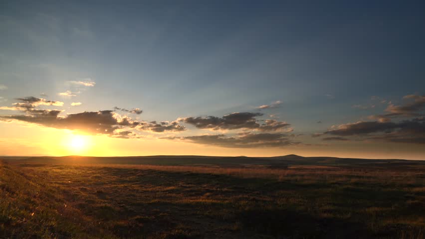 Mongolia sunset over treeless steppe and grassland beneath clouds across a vast empty plain. Golden evening sun lights an open prairie vista with barren fields, dusky heaven, no humans. Tracks, houses, fields, orchards, fences, corrals, grazing areas connect natural setting with everyday regional life. Human presence stays readable through settlement pattern, cultivated parcels, working infrastructure rather than staged activity. Pastoral details add social context, suggesting agriculture, remote habitation, seasonal labor, long established relationships with land. Open countryside spacing preserves authenticity, because buildings, routes sit naturally within surrounding relief, vegetation patterns. Layered relief, exposed strata, broken slopes, distant ridgelines give strong geographic structure across the entire view. Rock, soil, scrub, forest cover, open meadow patterns emphasize landform contrast shaped by climate, long erosion. Elevation change becomes especially clear where valleys, plateaus, cliffs, basins, high ridges organize visual rhythm across frame. Regional character feels convincing thanks to genuine topography, broad horizons, unforced relationships between surface texture, contour. Changing light, cloud build up, afterglow, rain haze, spectral color introduce time based drama without losing clarity. Atmospheric variation gives footage extra narrative range, from calm transition, renewal to threat, release, wonder. Sky behavior interacts with ground forms through shadow, brightness, color shift, enriching depth, seasonal expression. Passing weather adds realism, because transient illumination, moving vapor keep landscape visually alive. Foreground detail anchors scale, middle distance builds depth, remote horizon completes convincing spatial continuity. Natural color variation separates primary forms, secondary textures, background layers without artificial stylization. Additional visible cues suggest mongolia, sunset, steppe, grassland, sky, mongolian steppe.