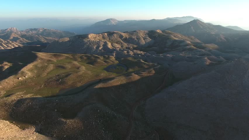Aerial view of the Atlas Mountains stretching across Algeria and North Africa in a semi-arid climate. Rugged peaks of the High Atlas and Anti-Atlas ranges form dramatic desert-steppe landscapes under clear skies. Layered relief, exposed strata, broken slopes, distant ridgelines give strong geographic structure across the entire view. Rock, soil, scrub, forest cover, open meadow patterns emphasize landform contrast shaped by climate, long erosion. Elevation change becomes especially clear where valleys, plateaus, cliffs, basins, high ridges organize visual rhythm across frame. Regional character feels convincing thanks to genuine topography, broad horizons, unforced relationships between surface texture, contour. Wide framing preserves spatial clarity, while motion direction keeps depth readable from foreground texture toward distant forms. Steady observation supports documentary realism, because composition stays clean, legible, easy to interpret during continuous movement. Panoramic sweep or fixed perspective provides usable establishing material for travel, geography, environment, atmospheric storytelling. Scale relationships remain strong, since vantage, horizon placement, gradual transitions reveal authentic proportions across terrain. Foreground detail anchors scale, middle distance builds depth, remote horizon completes convincing spatial continuity. Natural color variation separates primary forms, secondary textures, background layers without artificial stylization. Ambient mood remains consistent, giving footage polished yet realistic observational character. Lighting preserves tonal nuance, edge definition, material contrast instead of exaggerated treatment. Textural richness rewards slow viewing, because surfaces, silhouettes, motion cues remain easy to study. Authentic environmental cues help the scene read clearly for geography, ecology, climate, travel narratives. Additional visible cues suggest algeria, algerian, arid, morocco, moroccan, atlas mountains.
