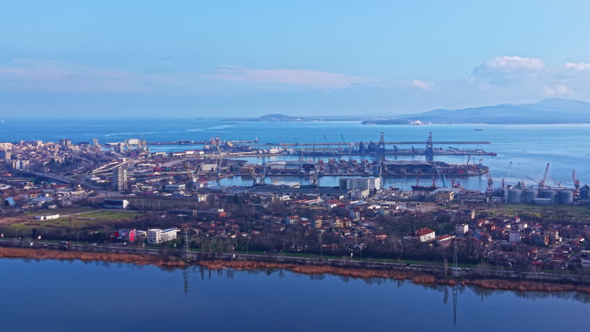 The scene shows a large industrial port with multiple cranes and ships docked at the shore. Calm waters reflect the structures under a bright blue sky.