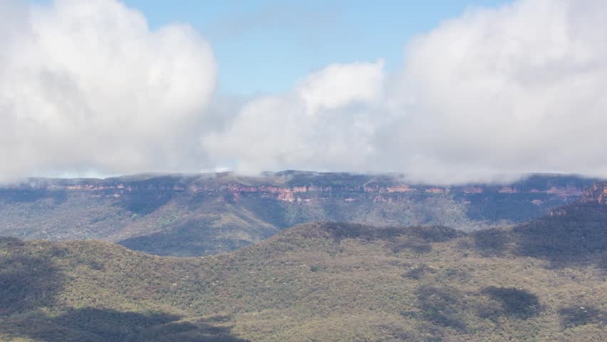 Time-lapse footage of a scenic highland landscape featuring dense green forested hills and a striking plateau with reddish-brown rock faces. Low-hanging clouds drift across the sky, casting shadows.