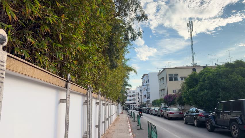 A street view in Rabat with a tall white fence and lush green bamboo. This urban scene captures the peaceful atmosphere of Morocco under a cloudy sky. Cars are parked along the road next to modern buildings and a telecommunication tower.