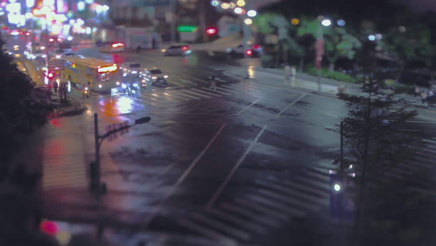Buses and cars navigate a wet Shilin Night Market intersection at night, captured in tilt-shift with glowing traffic reflections.